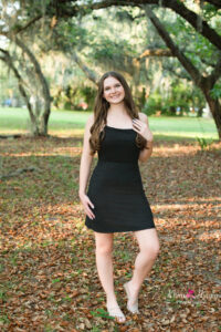 oviedo senior girl in a black dress is at lake mills standing up a shaded tree as khim higgins photography takes her photo.