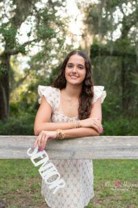Oviedo high school senior girl leaning against a fence holding a wooden SENIOR sign with Khim Higgins Photography.