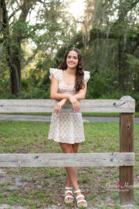 oviedo high school senior girl in a short floral dress is leaning on a wooden fence as Orlando senior photographer, khim higgins snaps her photos.