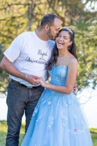 a sweet 16 girl wearing a blue lacy dress is standing next to her dad as he kisses her forehead.