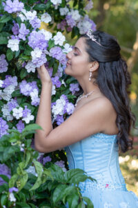 a sweet 16 girl in a blue dress is smelling purple flowers at Leu Gardens.
