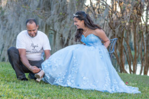 a sweet 16 girl in a blue dress is sitting on a chair as her dad puts on her shoes.