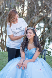 A sweet 16 girl in a blue dress is sitting on a chair while her mom puts a tiara on her head.
