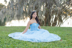 a sweet 16 girl in a blue dress is sitting on the lawn as Khim Higgins Photography takes her photos.