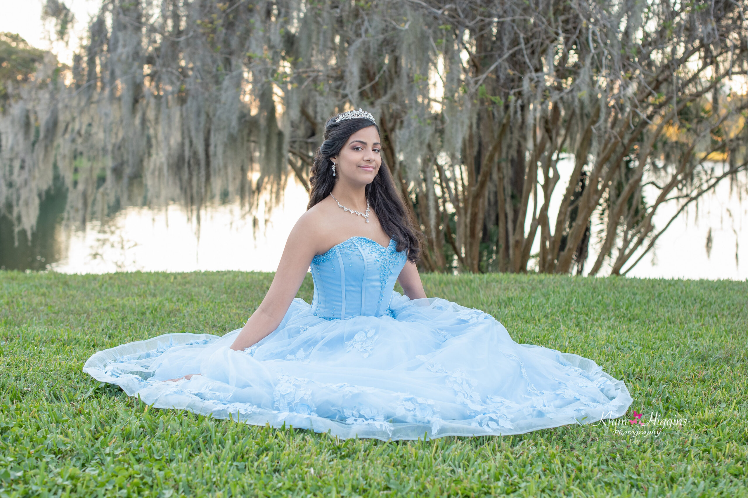 a sweet 16 girl in a blue dress is sitting on the lawn as Khim Higgins Photography takes her photos.