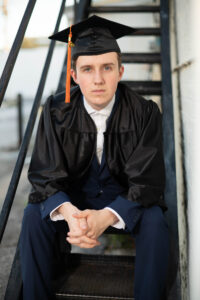 Senior boy wears a cap and gown sits on stairs.