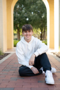 A senior boy sitting at a Rollins archway smiling for his senior photos with Khim Higgins Photography.